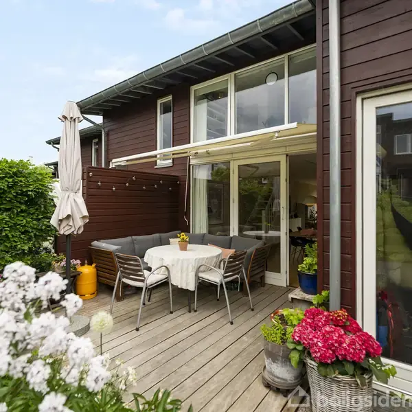 Terrasse med træbord og stole omgivet af blomster og planter. Et brunt træhus med store vinduer i baggrunden. Parasol foldet sammen ved siden af siddeområdet.
