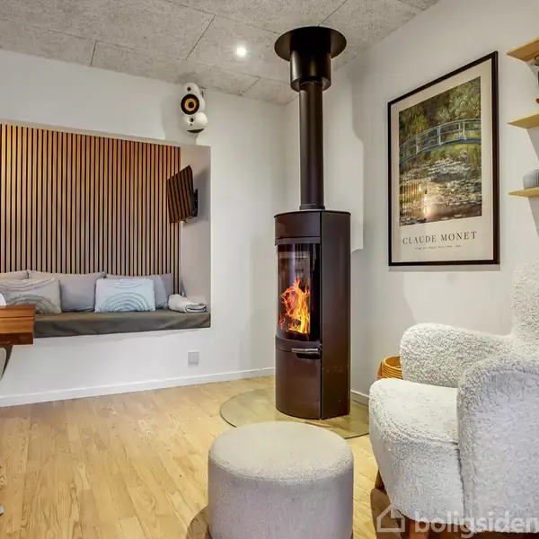 En brændeovn er tændt i et moderne stuehjørne med en hvid stol, vægkunst af Claude Monet og træhylder. I baggrunden ses en indbygget bænk med puder.
