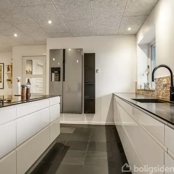 Køkken med hvide skabe og sorte bordplader ligger i et rum med grå klinker. Der er et stort køleskab for enden og en pejs i baggrunden.