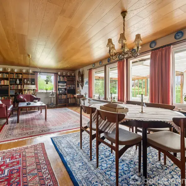 Spisestue med spisebord og stole, omgivet af bogreoler og tæpper. Store vinduer med røde gardiner giver udsigt til en terrasse og have. Lysekrone hænger fra træt loft.
