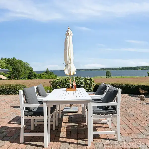 Et hvidt havebord med seks stole står på en flisebelagt terrasse. Parasol står i midten af bordet. I baggrunden ses grønt landskab og en sø under en blå himmel.