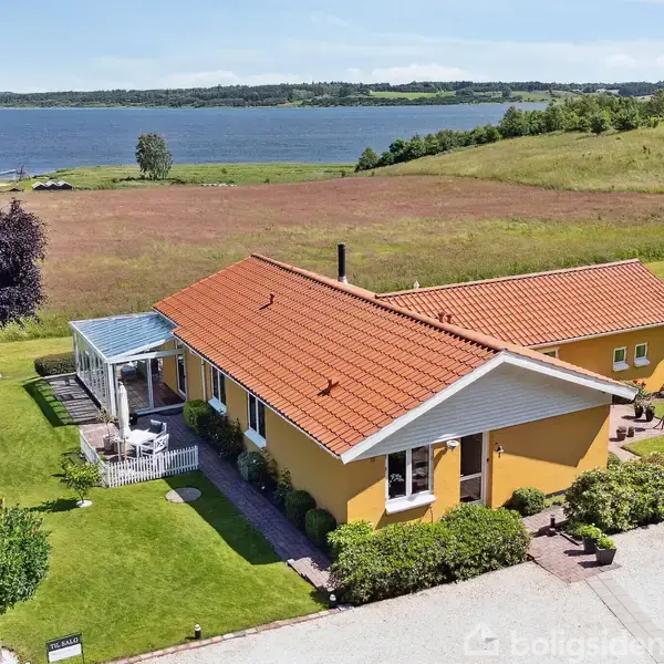 Hus med gult murværk og rødt tegltag står i en grøn have med en terrasse. Udsigt over en rolig sø og marker i baggrunden.
