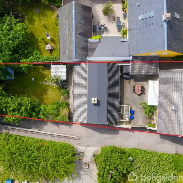 To bygninger set oppefra er adskilt af en mindre vej. Byområdet er omgivet af træer og grønne haver. En rød linje markerer en rektangulær ejendom.