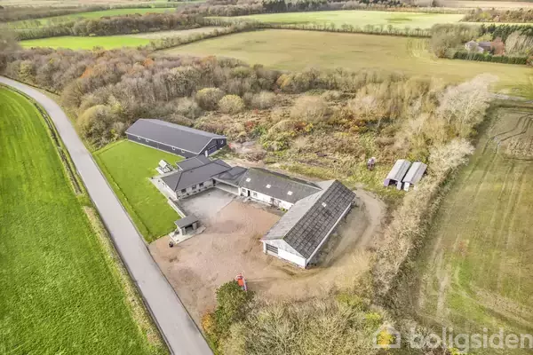 Gårdkompleks ligger roligt i landlig omgivelse med grønne marker og træer omkransende. Bygningerne inkluderer hovedhus og udhus, beliggende ved en asfalteret vej.