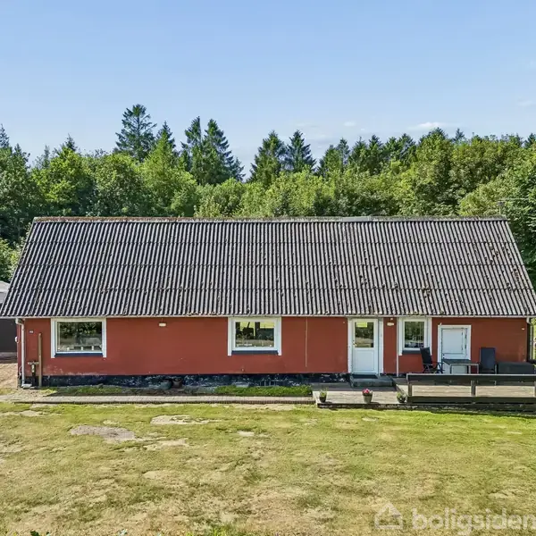 Rødt hus med sort tag, står stille, omgivet af grønne træer og græsplæne. Terrasse med stole foran. Sportsudstyr og opbevaringsskur til venstre. Klar, solrig dag.