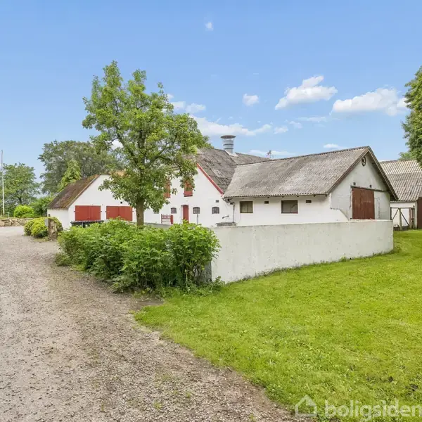 Et hvidt, stråtækt hus med røde vinduesrammer står omkranset af grønne træer og buske på en grusvej. Græsplæne på højre side, blå himmel i baggrunden.