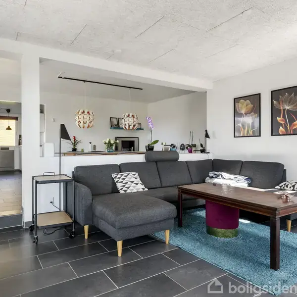 En grå sofa står i en stue med en træbord foran, dekoreret med en blå tæppe. Væggen har tre blomstermalerier. Rummet har et åbent design mod køkkenet, som ses i baggrunden.