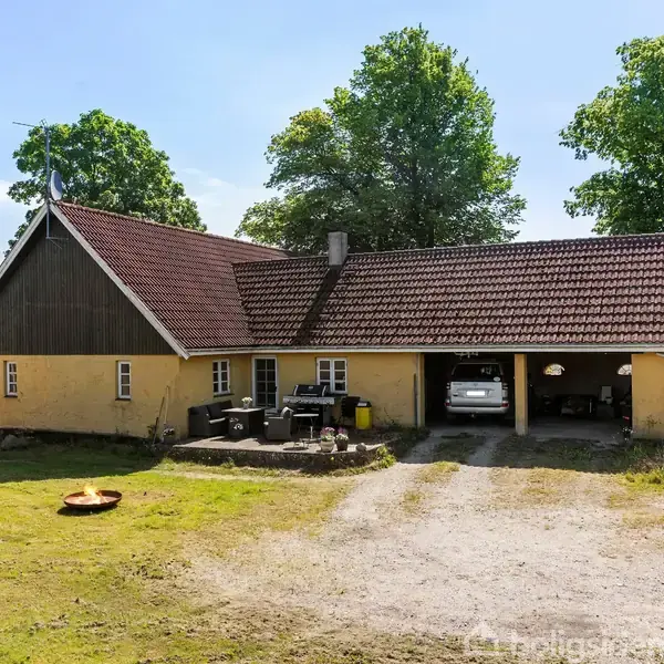 Gult hus med brunrødt tag står midt i en grøn have med store træer. En bil er delvist parkeret i en åbning. Grusvej fører op til huset.