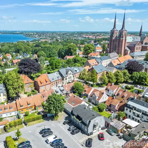 Roskilde Domkirke dominerer horisonten med to spir. Omgivet af tæt bebyggede røde tage og grønne træer, ses også parkerede biler på en parkeringsplads og en udsigt over vandet i baggrunden.