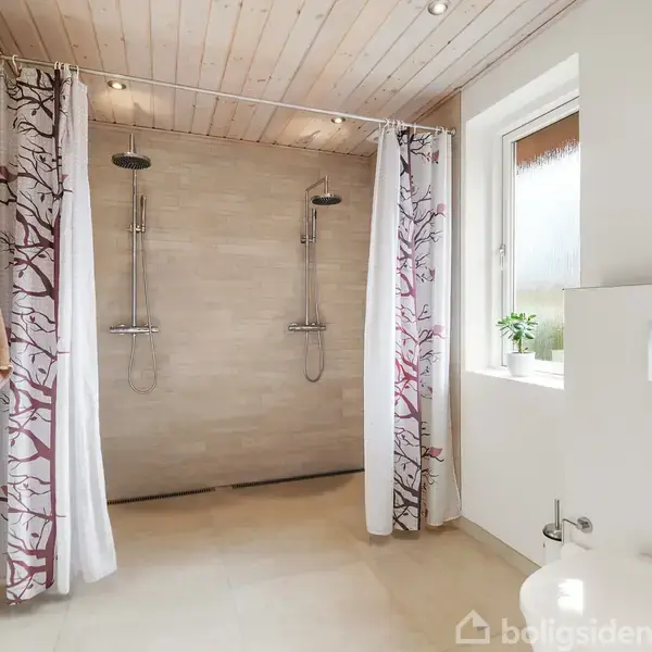 Dobbelte brusehoveder hænger på en væg i et lyst badeværelse. Bruseforhæng med træmotiv hænger delvist åbne. Et håndklæde hænger på en opvarmet ståltørrestativ ved siden af. En lille plante står ved vinduet.