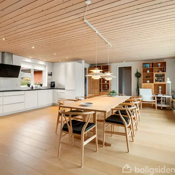 Et moderne køkken-alrum med et stort træbord og stole, åben til et lyst rum med store vinduer, integreret køkken med hvide skabe og apparater, trægulv og loftsbelysning.