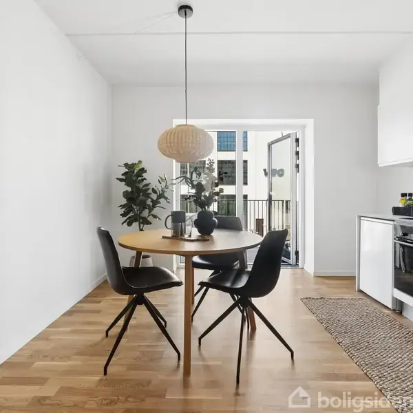 Rundt spisebord med tre stole står i et lyst køkkenalrum. Bordet har planter og dekorationer. Til højre er der et køkken med hvidt skab og komfur. Store vinduer i baggrunden.