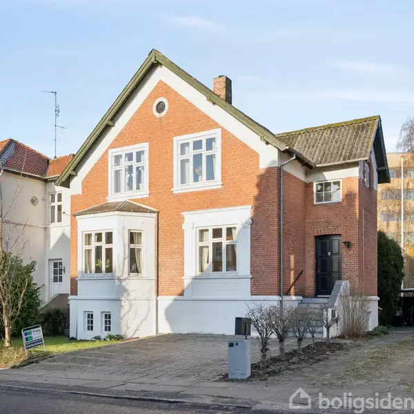 Rødt murstenshus med hvidmalede kanter står på en flisebelagt indkørsel. Det har et sort skråt tag og omfavnes af tætstående nabohuse og træer i en beboelsesområde.