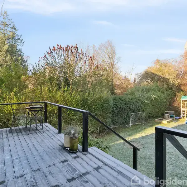 En træterrasse har en stol og planter; den leder ned til en frodig, let frossen have med legeredskaber og buske. En frostklar himmel omslutter baggrunden.