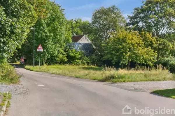 En vej omgivet af grønne træer og buske fører til et hus i det fjerne. Trafikskilte advarer om ujævn vejbane og indkørsel forbudt.