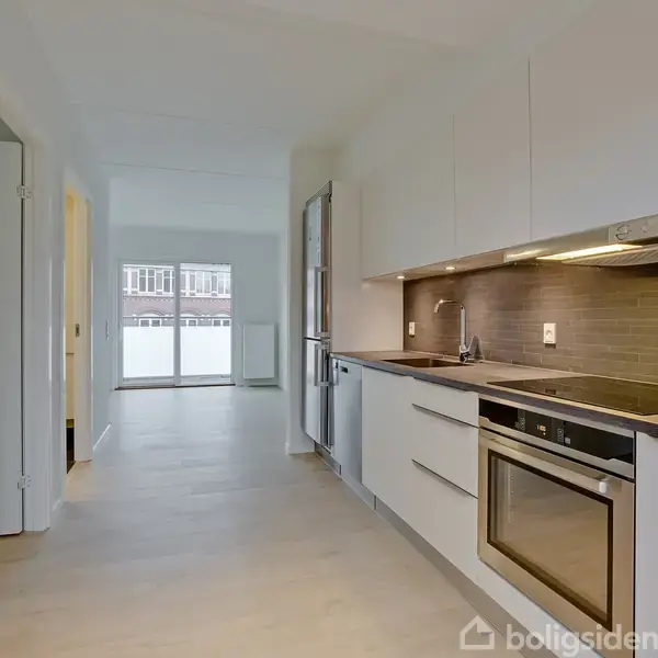 Et lyst køkken med hvide skabe og indbygget ovn; ligger i et åbent rum med lys gennem store vinduer i baggrunden. Moderne inventar og træfarvede fliser fuldender indretningen.