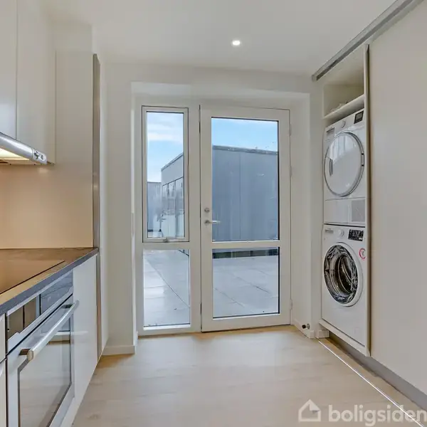 En moderne vaskemaskine og tørretumbler er stablet i et skabsområde ved siden af en køkkenbænk med ovn og kogeplader. I baggrunden ses en glasdør mod terrassen.