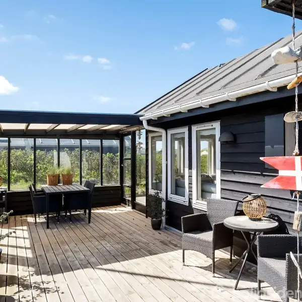 Sort træhus med stor træterrasse. Et bord med stole er placeret på terrassen, der er omkranset af glasværn. En lille rød flagdeko hænger fra taget. Solrigt og grønt område.