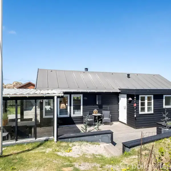Sortmalet hus med terrasse og tilhørende udestue i solrigt område, omgivet af græsplæne og enkelte buske. Blå himmel og sollys præger omgivelserne, hvilket giver et lyst og åbent miljø.