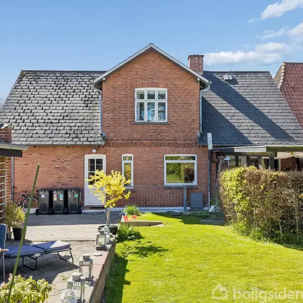 Murstenshus med sort skifertag, stående stille i solrigt kvarter. Foran er en velplejet grøn have og en terrasse med møbler, skraldespande på siden. Blå himmel i baggrunden.