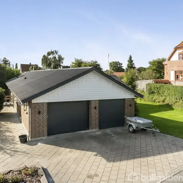 Garage med to porte står stille på brolagt indkørsel. Ved siden af ses en lille trailer. I baggrunden er der en stor græsplæne og et rødt hus med have.