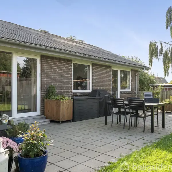Bygning i mursten med store vinduer. Terrasse med fliser, havemøbler og planter i potter. Haven er grøn og omgivet af hegn. En stor hængepil træ til venstre.