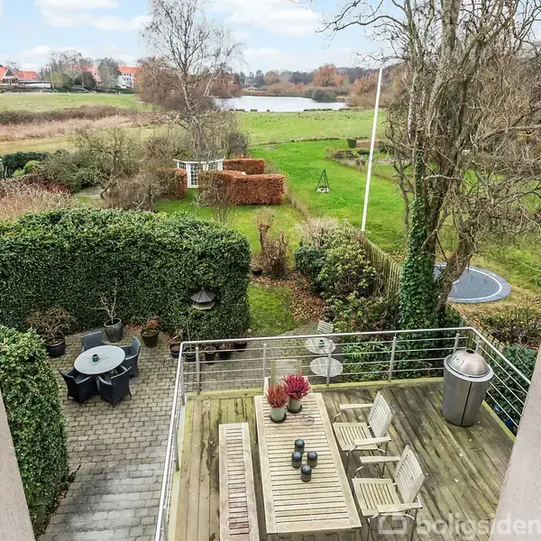 Terrassebord med stole står på træterrasse. Rundt bord i sten på flisebelagt område med planter. Græsplæne med sø og buske i baggrunden. Trampolin ses blandt træerne.