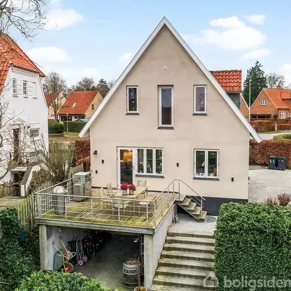Et hvidt hus med en spids gavl har en stor terrasse foran. Der er en trappe, der fører op til terrassen. Huset står i et villakvarter med andre huse.