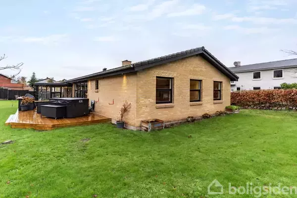 Et gulstenshus med fladt tag står på en velplejet græsplæne. Der er en træterrasse til venstre med en stor, udendørs jacuzzi. I baggrunden ses andre boliger.