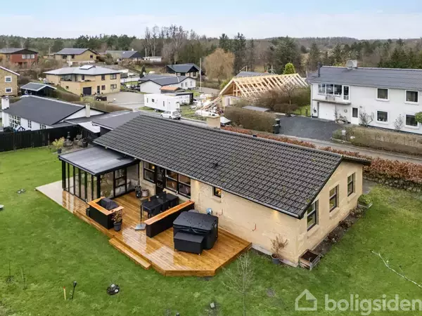 Et enfamiliehus med fladt tag ligger på en grøn græsplæne. Huset har en stor træterrasse med spabad. Omkringliggende huse og et kvarter ses i baggrunden.