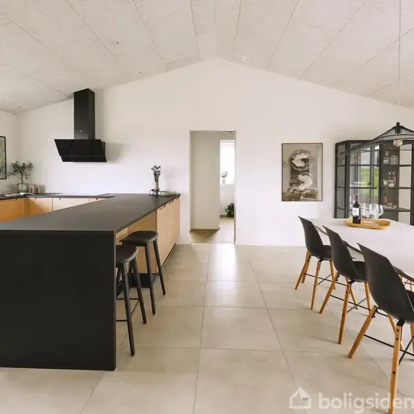 Køkken- og spiseområde med lang sort køkkenø, barstole, og træpaneler, åben til spisestue med stort hvidt bord, sorte stole, og vitrineskab. Flisegulv og hvidt loft i moderne design.