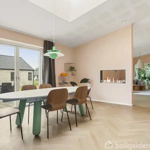 Spisebord med seks stole står i et lyst rum med lyserøde vægge og skråloft. Et vindue med gardiner viser en udsigt til et hus tag. En moderne pejs pryder væggen.