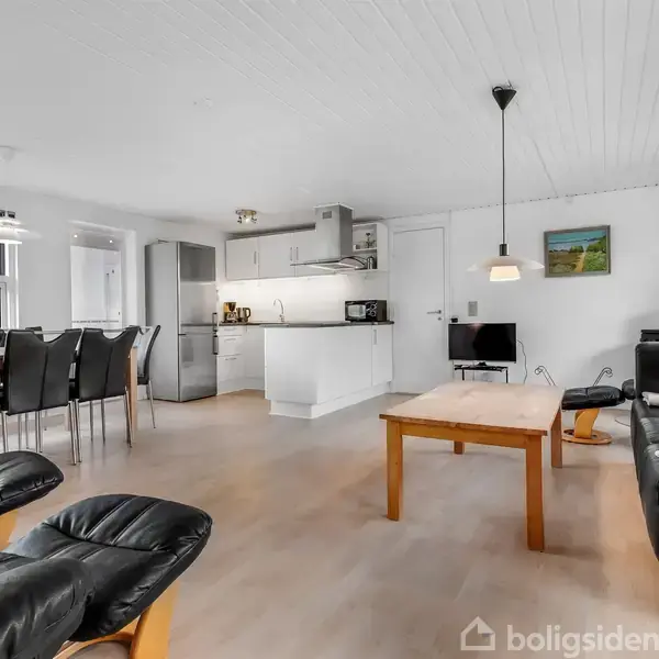 Stue indrettet med sort lædermøbler, spisebord med sorte stole til venstre, og et køkkenområdet i baggrunden. Et TV står på en lille stander, og en lampe hænger fra loftet.