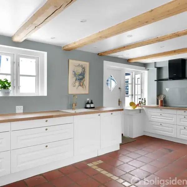 Køkken med hvide skabe og træbordplade, indeholder ovn og komfur. Vægdekorationer og planter ved vinduet. Loftet har synlige bjælker. Gulvet er af rødlige fliser med lysindfald fra flere vinduer.