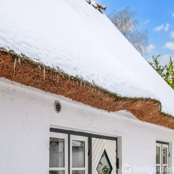 Et stråtækt hus med isdannelse på taget under sneen. Huset har hvide vægge og små vinduer. Omgivet af grønne træer og klar blå himmel med skyer.