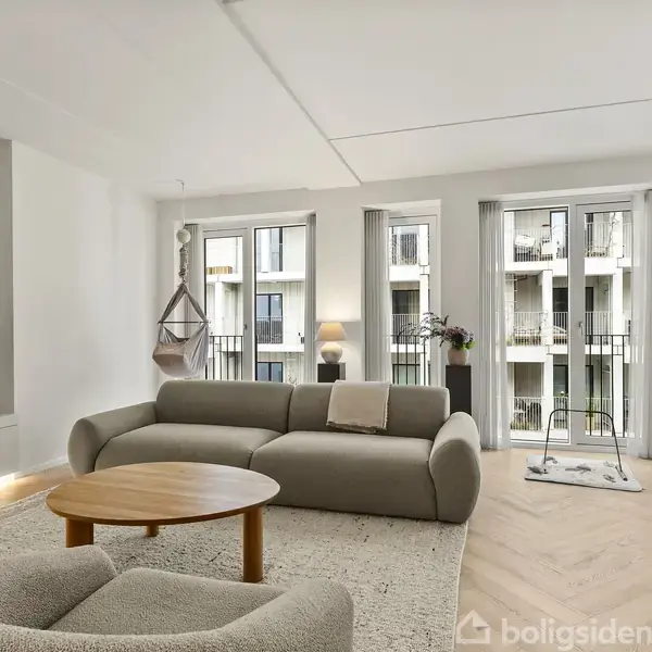 Grå sofa og rundt træbord står i en lys stue med tæppe, fladskærms-TV på væggen og store vinduer med udsigt til altaner.