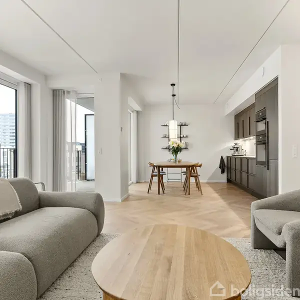 Grå sofaer står i en stue ved siden af et trægulv, hvor et spisebord og stole er placeret. Køkkenet er i baggrunden, og store vinduer giver naturligt lys.