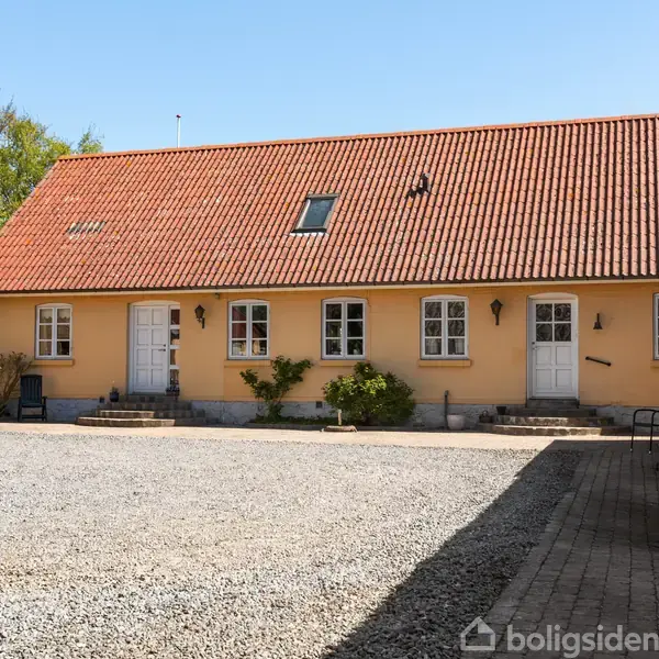 Gult murstenshus med rødt tegltag, døre og vinduer i facaden. Gruset gårdsplads foran, omkranset af grønne træer i baggrunden.