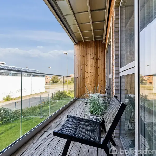 Balkon med sorte stole og planter i potter langs glasrækværk. Sollys skinner ind fra venstre og afslører en bymæssig baggrund med bygninger og en vej.