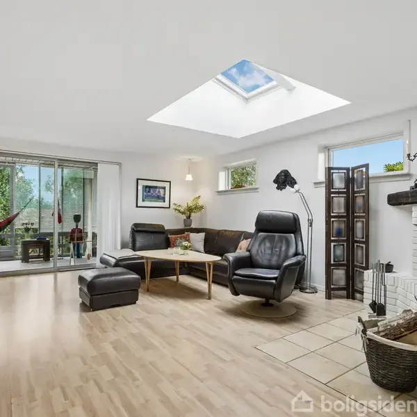 Stue med moderne møbler, herunder lænestole og sofabord, står foran pejsen. Store vinduer og ovenlysvindue giver rummet lys, mens terrassedør åbner mod grøn udsigt.