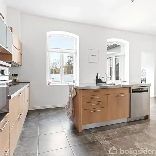 Et moderne køkken med lyse træskabe og en central ø, der har integreret opvaskemaskine. To store vinduer giver naturligt lys. Et skilt på væggen siger "California".