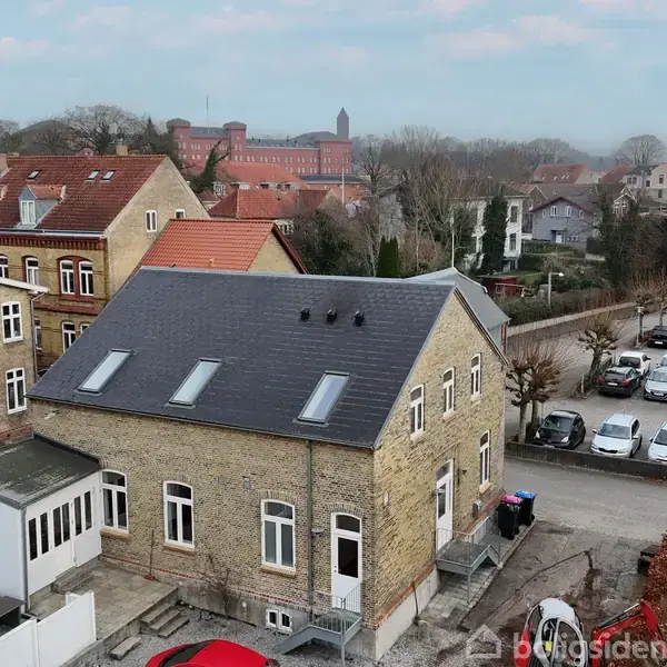 Et gulstenshus med sort skråtag står i et villakvarter. Til venstre er et lignende hus. Flere biler er parkeret langs en allé bag huset. I baggrunden er der flere bygninger.