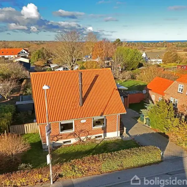 Et rødt murstenshus med orange tegltag står stille i en villavej. Det er omgivet af haver og andre huse under en blå himmel med spredte skyer.