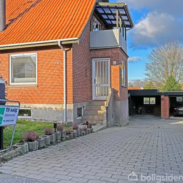 Rødt murstenshus med orange tag, trappe ved hoveddør. Skilt med teksten "Til salg" fra Hybolig Ejendom ved indkørsel. Garage i baggrunden, blå himmel og træer omkring.