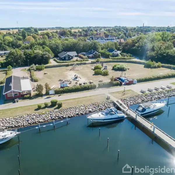 Flere lystbåde ligger forankret ved en bro langs en rolig kanal. I baggrunden ses et grønt område med huse og plæner under en klar blå himmel.
