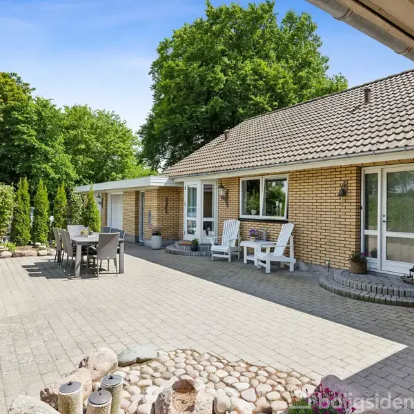 En gul murstensvilla med skråt tag står stille omgivet af en flisebelagt terrasse. Havemøbler og planter dekorerer området, med tæt, grøn vegetation i baggrunden.