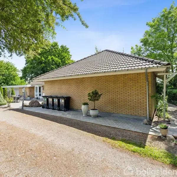 Et gult murstenshus med skråt tegltag står ved en grussti omgivet af grønt løv. Flere affaldsbeholdere og planter i krukker ses foran huset.