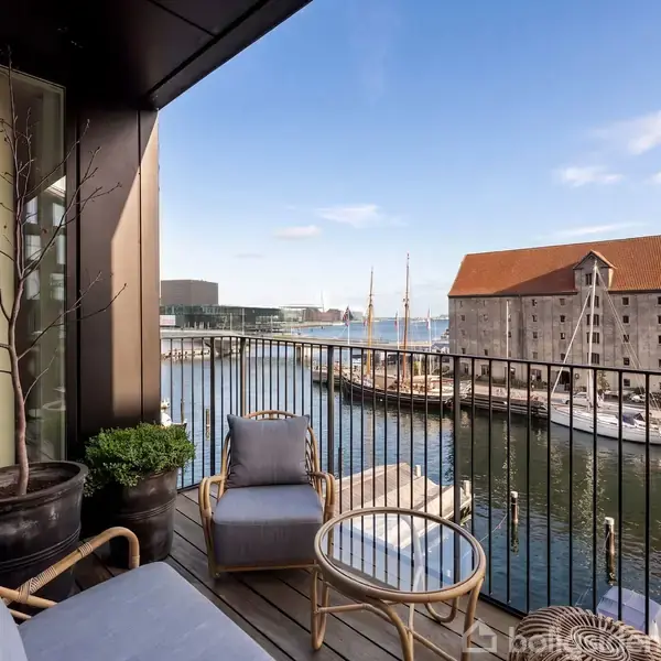 En altan med to stole og et bord tilbyder udsigt over en havn med et skib og en historisk bygning i baggrunden. Solrig himmel og afslappet atmosfære.