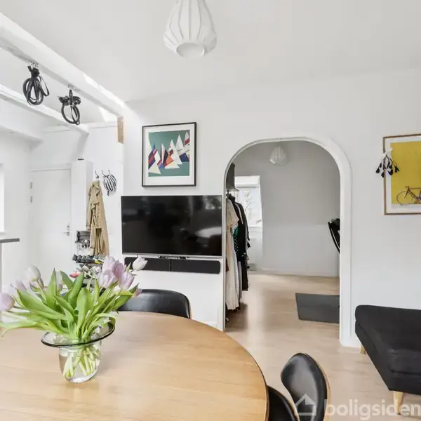 Rundt spisebord med vase af lyserøde tulipaner. TV står foran en hvid væg; cykelplakat og reol med bøger i rummet. Lys vælter ind fra store vinduer.