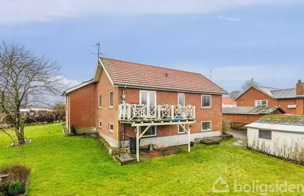 Rødt murstenshus med en balkon, beliggende i en græsbevokset have. En garage står til højre, og himlen er overvejende skyet udenfor. Der er ingen tekst i billedet.